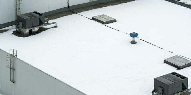 Snow-covered white rooftop of a building with HVAC units, skylights, and a blue vent. A black cable runs across the roof.