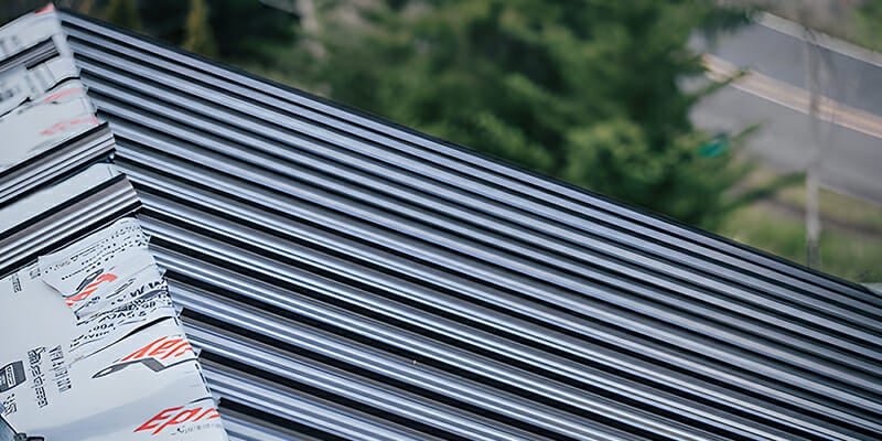 Close-up of a stack of metal roof panels on a roof, with blurred trees and road in the background.