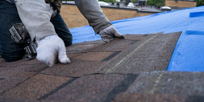 A roofer in gloves laying asphalt shingles on a roof partially covered with blue underlayment.